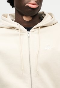 Φούτερ με φερμουάρ σε εκρού χρώμα και ομαλή υφή, με μικρό λευκό λογότυπο Nike στο αριστερό μέρος του στήθους και κορδόνια στη κουκούλα.