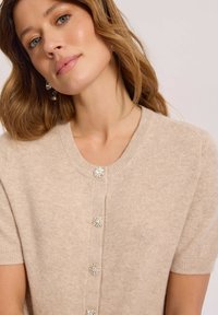 Kobieta z jasnobrązowymi włosami, nosząca beżowy kardigan z krótkim rękawem ozdobiony kryształowymi guziczkami oraz srebrne kolczyki.