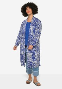 Lange, open voorkimono in een blauwe en witte abstracte print, gecombineerd met een blauwe top en cropped jeans, gedragen met grijze slide sandalen.