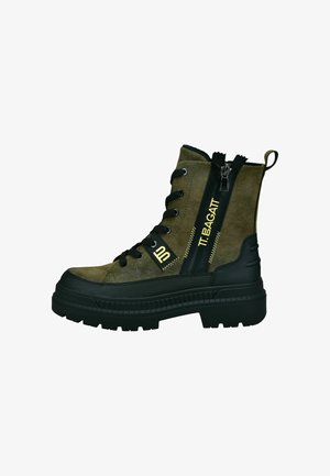 Stivaletto in pelle scamosciata verde oliva con suola in gomma nera, dotato di una cerniera laterale e lacci decorativi. Accento di branding giallo lungo il lato.