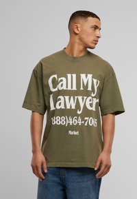 Oliven grøn bomulds t-shirt med korte ærmer, der har fed hvid tekst: "Ring til min advokat (888)464-7065" og "Marked" nedenunder.
