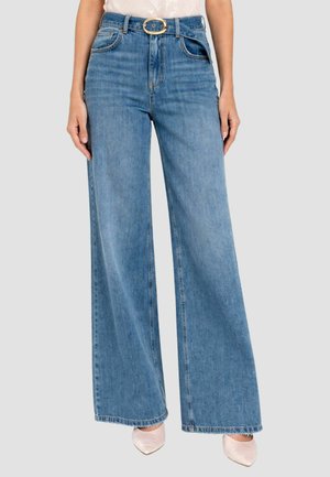 Jeans blu a vita alta con gamba larga e cintura con fibbia ovale dorata, indossati con scarpe a punta rosa pallido su sfondo neutro.