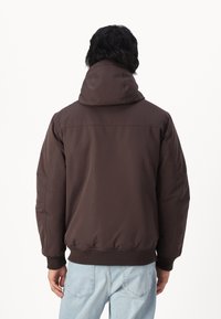 Brązowa kurtka z kapturem z wzmocnionym dołem i mankietami, o gładkiej fakturze i casualowym kroju. Noszona na jasnoniebieskich dżinsach.