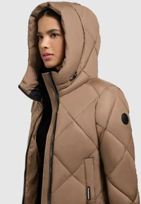 Beige vuorellinen puffer-takki, jossa on suuri huppu, vetoketjukiinnitys edessä ja logopatchi hihassa. Ominaisuudet sileä pinta ja timanttimallikuva.