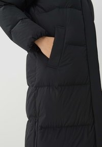 Zwarte pufferjack met een gestructureerd ontwerp, voorzien van een zijzak en zichtbare stiksels. Gladde, geïsoleerde textuur.