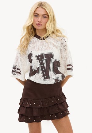 Femme blonde porte un top oversize en dentelle blanche avec des imprimés de lettres noires et une mini-jupe noire à volants avec des clous argentés.