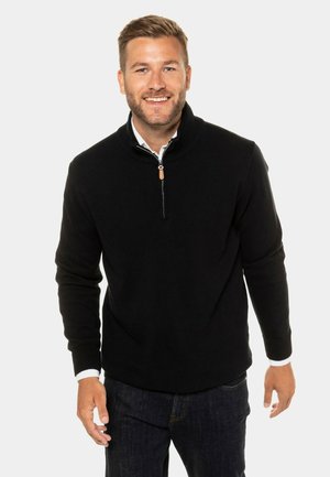 Schwarzer Strickpullover mit hohem Kragen und Reißverschluss, ausgestattet mit gerippten Bündchen und Saum. Kombiniert mit dunklen Jeans.