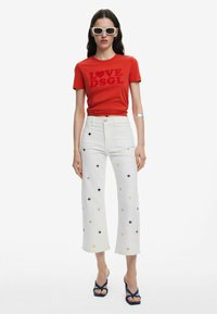 Rotes Baumwoll-T-Shirt mit dem Aufdruck "LOVE DSGL", kombiniert mit weißen weit geschnittenen Jeans, die mehrfarbige Blumenstickereien aufweisen. Blaue Sandalen.