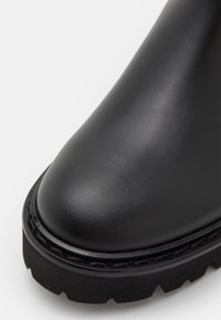 Bottine en cuir noir avec un bout arrondi, une surface texturée et une semelle épaisse avec des détails de motif de crampons. Couture détaillée le long du bord.