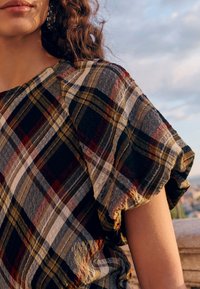 Camicia a quadri con maniche corte arruffate, realizzata in un tessuto strutturato con colori che includono verde scuro, bordeaux e accenti bianchi.