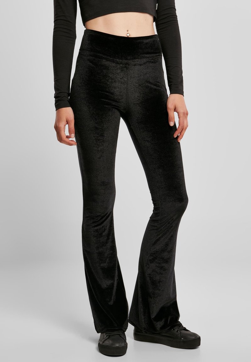 Personne portant un pantalon évasé en velours noir taille haute, des baskets noires à plateforme et un crop top noir à manches longues, debout sur un fond uni.