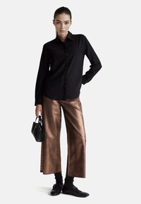 Czarna koszula na guziki, miedziane metaliczne spodnie o szerokich nogawkach, czarna torebka o fakturze oraz czarne buty z koronkowymi skarpetkami, na białym tle.