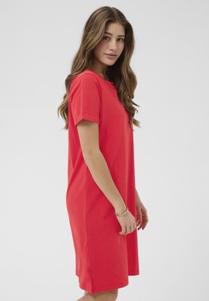 Junge Frau mit langen, welligen Haaren, die ein rotes, kurzärmeliges knielanges Kleid trägt und seitlich vor einem schlichten grauen Hintergrund steht.