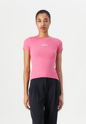Rosa geripptes Kurzarmoberteil mit einer engen Passform und einem weißen Logo auf der Brust, kombiniert mit dunklen, maßgeschneiderten Hosen.