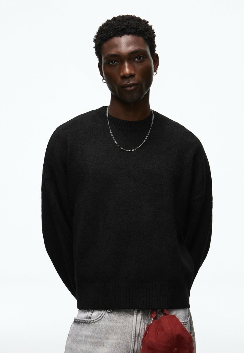 Jeune homme aux cheveux courts et bouclés porte un pull noir, un collier chaîne argenté, des boucles d'oreilles et un jean clair, tenant une casquette rouge derrière son dos.