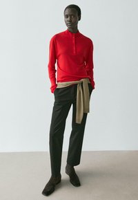 Rotes Langarmoberteil mit Knopfleiste, kombiniert mit schwarzen, zulaufenden Hosen. Ein beigefarbener, gestrickter Pullover ist um die Taille gebunden. Braune Stiefeletten.