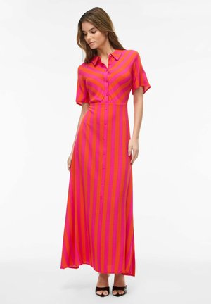 Kvinne som har på seg en gulvlengde, knappet kjole med vertikale røde og rosa striper, korte ermer og svarte åpne tå-hel sko, står mot hvit bakgrunn.