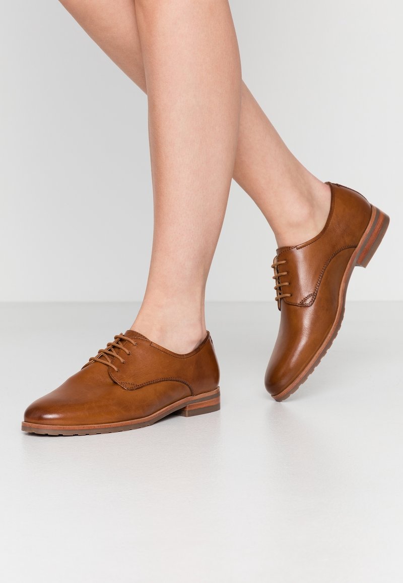 Femme portant des chaussures Oxford en cuir marron à lacets, debout sur un sol gris clair avec un pied légèrement relevé.