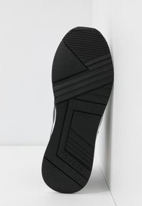 Semelle d'une basket noire avec des motifs géométriques de traction et la marque "GUESS" estampillée près du talon, posée contre un coin blanc.