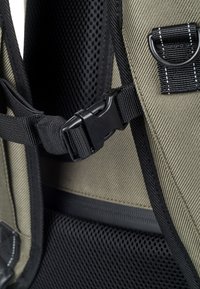 Khaki fabric backpack featuring black mesh padding, adjustable straps, a plastic buckle, and contrasting stitching details for durability.