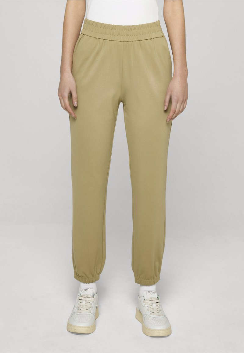 Személy bézs elasztikus derékpántos edzőnadrágot és fehér tornacipőt viselve, egy egyszerű világos háttér előtt áll.