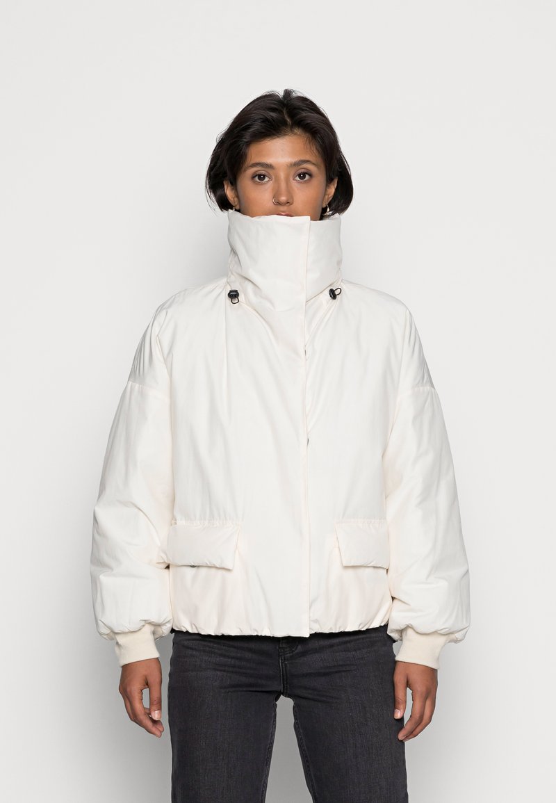 Veste blanche rembourrée à col montant, dotée de deux poches avant, d'une fermeture éclair et de poignets élastiques. Texture de tissu lisse.