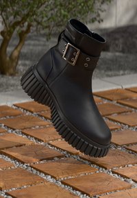 Crni gležnjača s debelim rebrastim potplatom i srebrnom kopčom, izložena na otvorenom na drvenom bloku i kamenčićima.