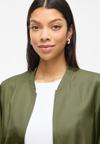 Olivovo zelená bomber bunda s hladkým, matným povrchom, so zipsom a rebrovaným golierom, nosená cez biely top.