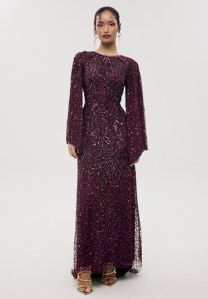 Femme portant une robe longue au sol, manches longues, violet foncé ornée de perles avec des motifs floraux et des talons ouverts dorés, debout devant un fond uni.