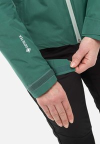Veste Gore-Tex verte avec ajustement des poignets par Velcro. Caractéristiques d'une fermeture éclair à l'avant et d'une texture de tissu lisse. Pantalons noirs visibles sous le poignet.
