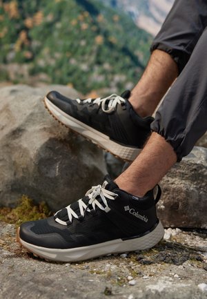 Czarne buty sportowe z białymi sznurowadłami, z teksturowaną siateczkową cholewką, gumową podeszwą i logo Columbia, umieszczone na skalistym terenie.