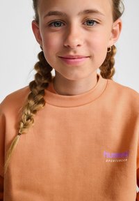 Pomarańczowa, prążkowana bluza z wysokim dekoltem, z fioletowym logo z napisem "hummel SPORTSWEAR". Modelka ma warkocze.