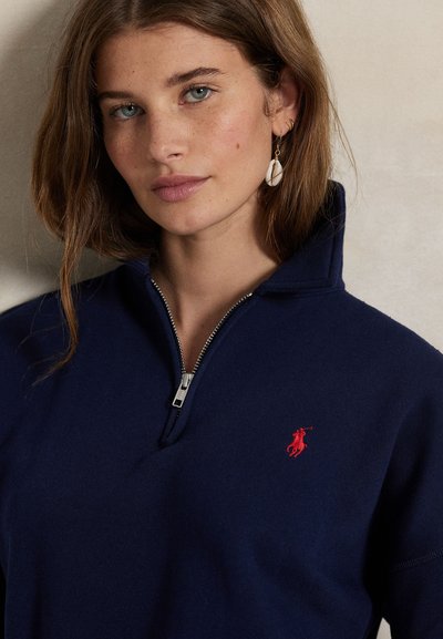 Mujer joven con cabello castaño claro, lleva un suéter azul marino con cremallera y logo bordado en rojo, y pendientes dorados en forma de concha, sobre un fondo neutro.