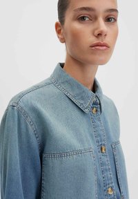 Camicia in denim blu chiaro con collo a apertura, chiusura a bottoni e due tasche sul petto con bottoni beige. Tessuto morbido e strutturato.