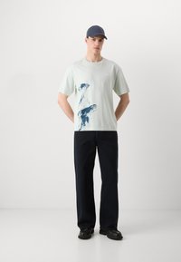 Lys grøn t-skjorte i bomull med abstrakt marineblå design, sammen med mørk marineblå bukser. Modellen har på seg en mørk blå caps og svarte sko.