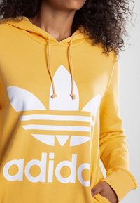 Gul hoodie med en vit Adidas-logotyp med tre ränder och ett treklövermönster på framsidan. Lättviktigt tyg med huva och dragsko.