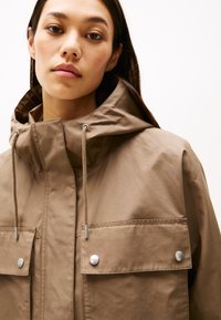 Brown waterproof jacket with a high collar, adjustable drawstrings, and silver snap buttons on two flap pockets. Smooth texture, minimal design.