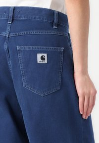 Džínsy šortky v tmavomodrej farbe s klasickým dizajnom piatich vreciek, obsahujúce zadné vrecko so štvorcovou nálepkou zobrazujúcou logo Carhartt.