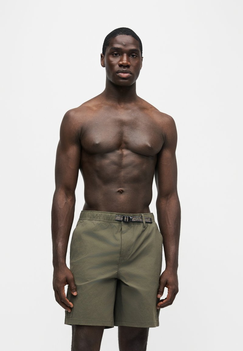 Homme en forme à la peau foncée debout, portant un short vert avec une ceinture ajustable noire et grise intégrée, sur un fond blanc uni.