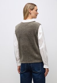 Grön fluffig väst med rundad halsringning, bärs över en vit blus. Västen har en kortad design och kombineras med blå denimjeans.