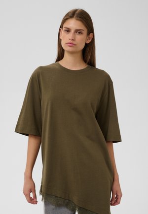 Jeune femme aux cheveux bruns et raides portant un t-shirt oversize vert olive avec une bordure en dentelle à l'ourlet sur un fond uni.