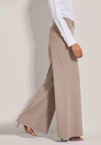 Beige wijde broek gemaakt van soepel materiaal, met een hoge taille en achterzakken, gecombineerd met strappy hoge hakken.