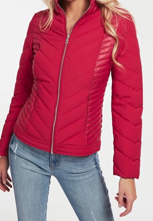 Mujer con chaqueta acolchada roja ajustada con cremallera frontal y jeans azules claros, con las manos relajadas a los lados contra un fondo liso.