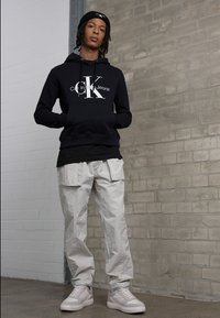 Crna kapuljača s bijelim logotipom "Calvin Klein Jeans", uparena s bijelim cargo hlačama i svijetlim tenisicama na teksturiranoj sivoj pozadini.