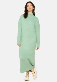 Minta zelena pletena obleka z visokim ovratnikom, dolgimi rokavi in sproščenim krojem, ki ima rebraste manšete in stranski razporek za udobno gibanje.