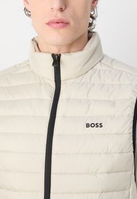 Beige prošívaná vesta s vysokým límcem, černým zipem a vodorovným prošitím. Na levé části hrudi je umístěno černé logo.