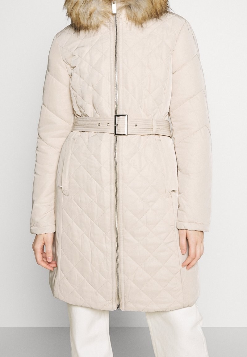Manteau matelassé beige avec boucle de ceinture, fermeture éclair avant, poches latérales, manches longues et col en fausse fourrure, porté avec un pantalon blanc.