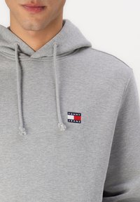 Hanorac gri din bumbac, cu buzunar tip cangur și glugă ajustabilă cu șnur. Prezintă un mic logo "TOMMY JEANS" pe piept.