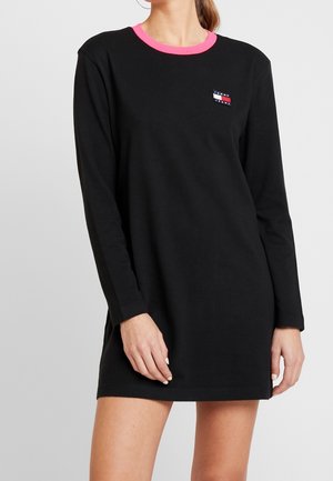 Kvinde iført en sort langærmet kjole med en pink krave og et lille Tommy Jeans-logo på brystet, stående mod en ensfarvet baggrund.