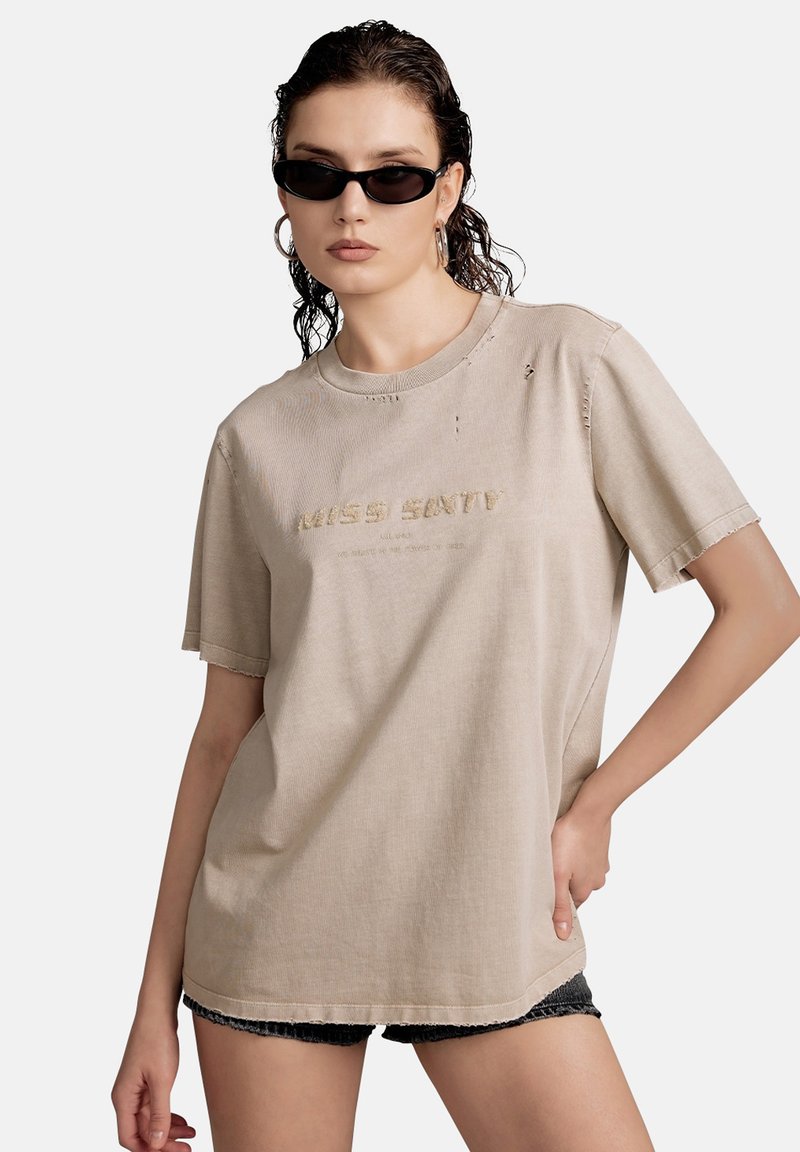 Camiseta oversized beige de tejido suave con detalles desgastados y texto empotrado "MISS ENTITY". Manga corta y escote redondo.